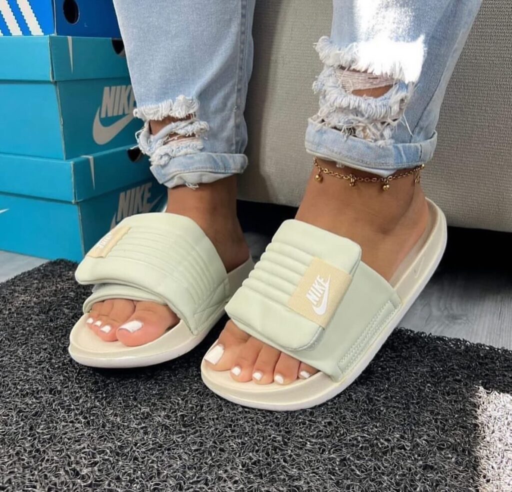 Light green Nike slide sandals worn with ripped blue jeans on a grey mat.