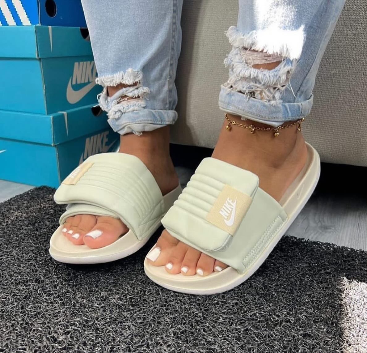 Light green Nike slide sandals worn with ripped blue jeans on a grey mat.