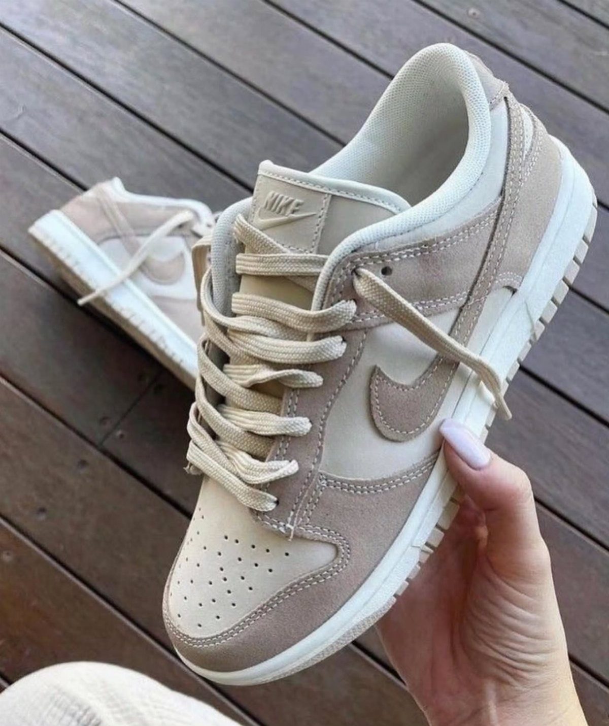 Beige and tan Nike Dunk sneakers held by a hand over wooden decking.
