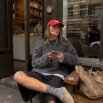Woman wearing tan Birkenstock clogs, grey sweater, and red cap sitting outdoors.