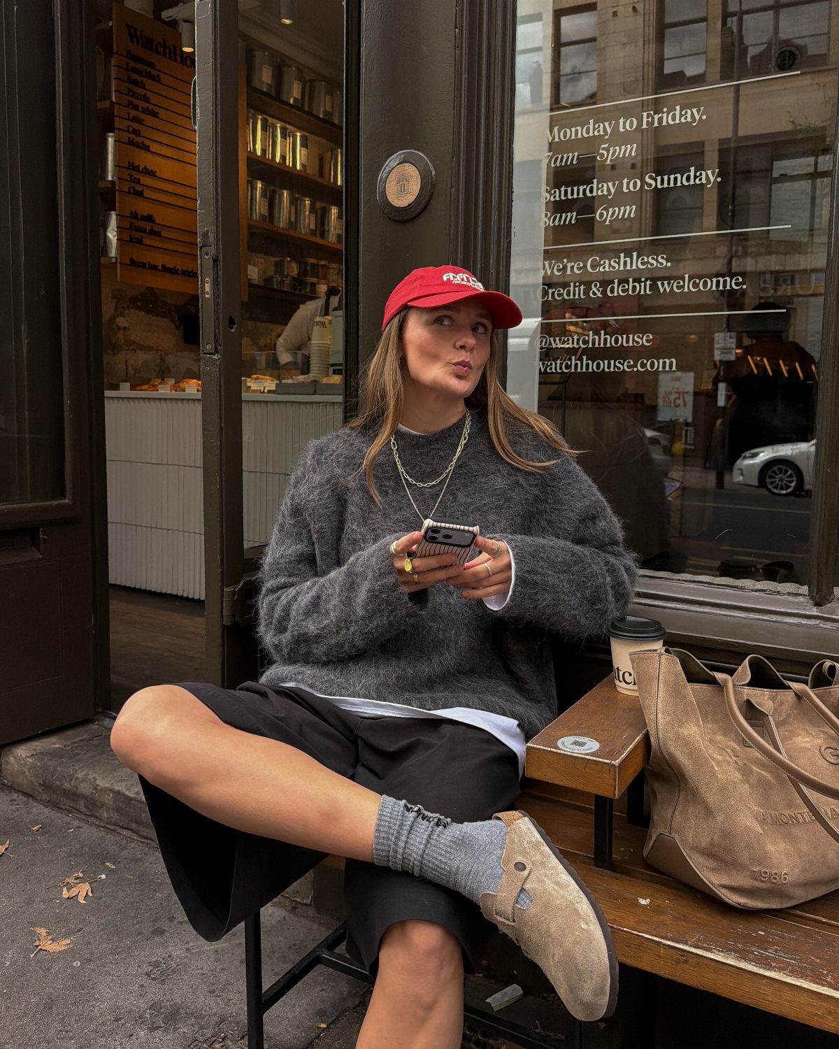 Woman wearing tan Birkenstock clogs, grey sweater, and red cap sitting outdoors.