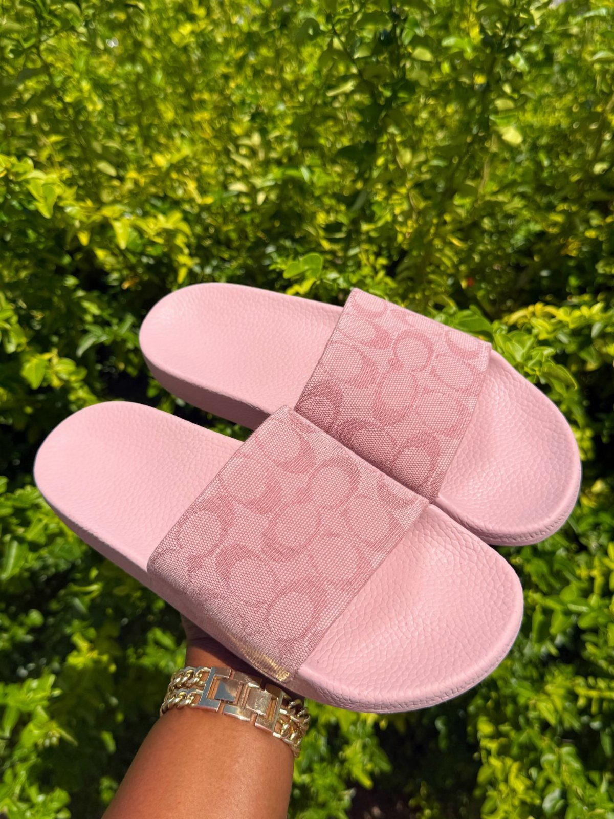 Pink slide sandals with logo straps held against a lush green leafy background.