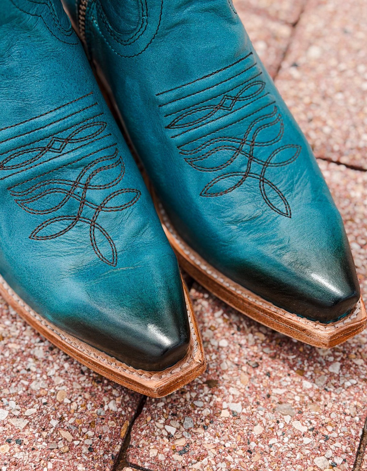 Teal leather cowboy boots with black decorative stitching on a stone pavement