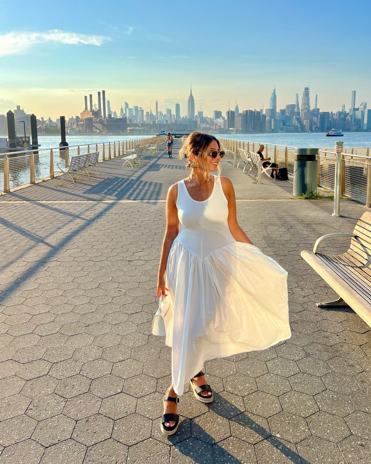 Woman in a white drop-waist dress standing on a waterfront with city skyline.
