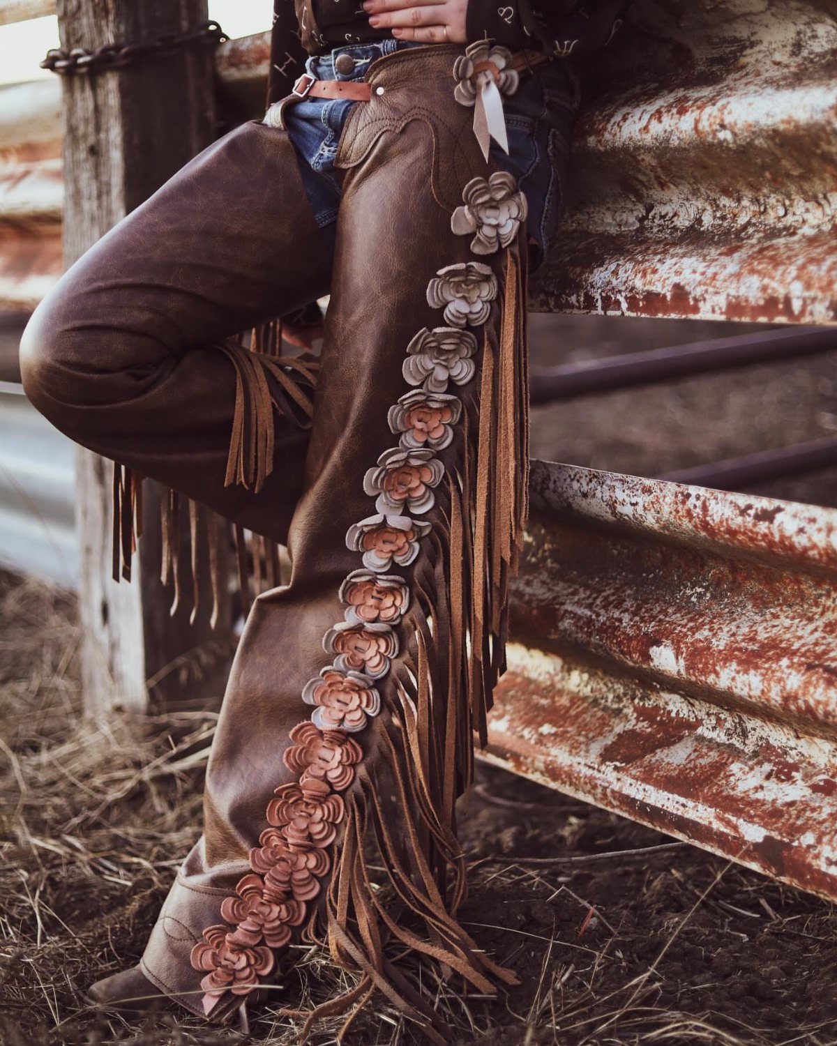 Brown leather chaps with floral carvings and fringe worn with blue jeans.