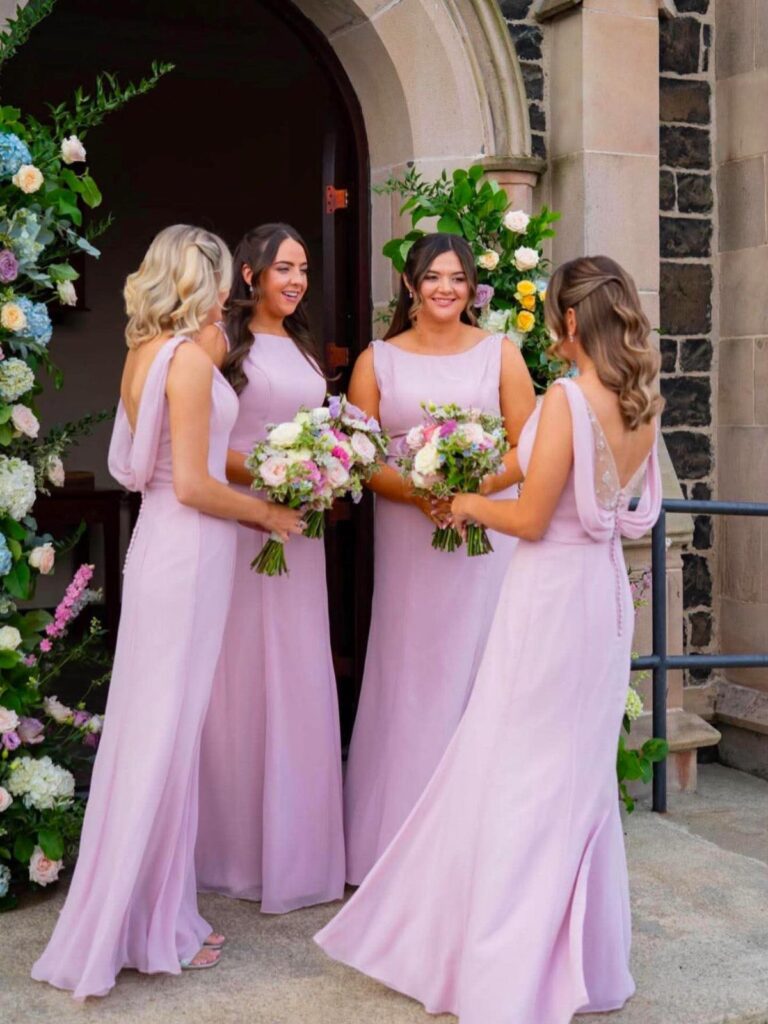 Four bridesmaids in light purple dresses holding floral bouquets outside a stone building.