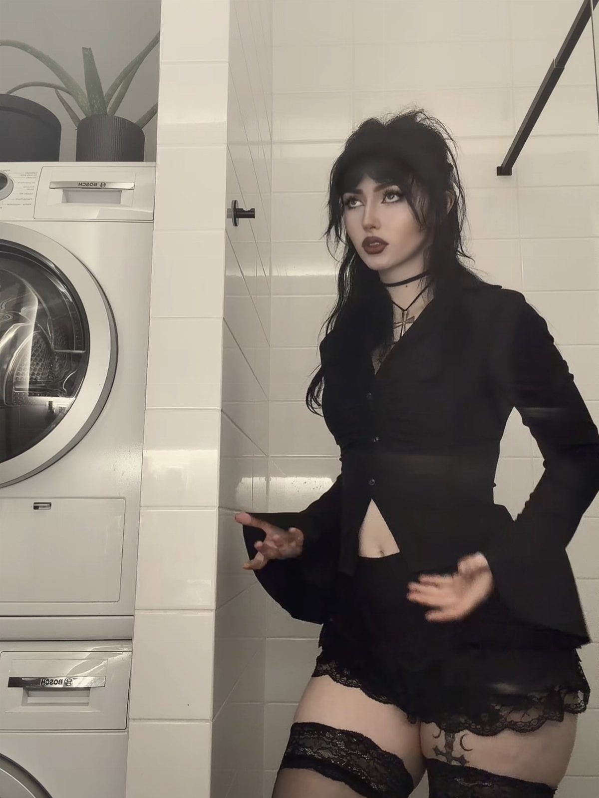 Woman in a black goth chic outfit posing in a white tiled laundry room.