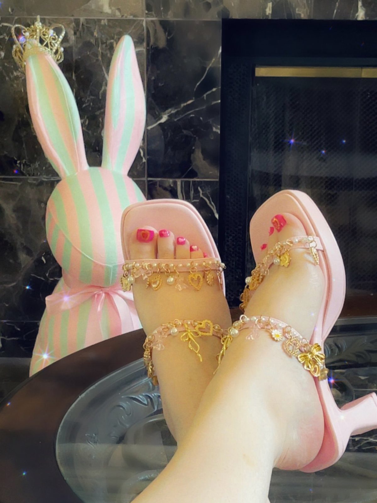 Pink high-heeled sandals with gold charms and beads on a person's feet.