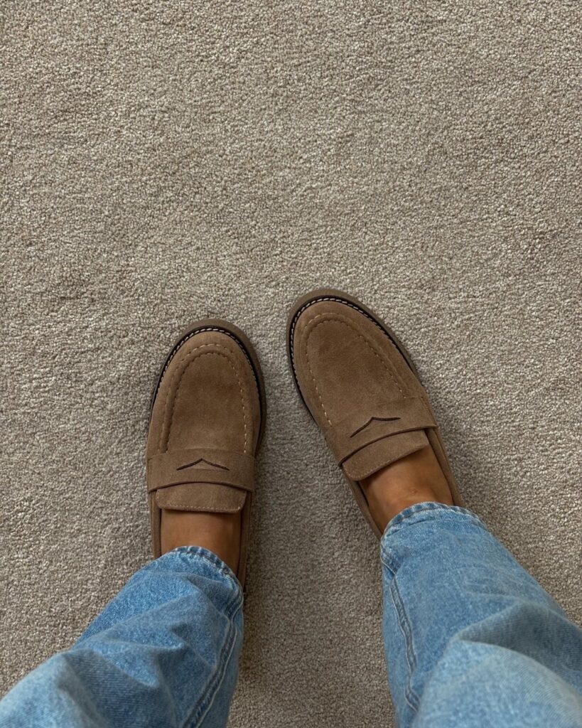Tan suede loafers worn with light blue jeans on a beige carpet.