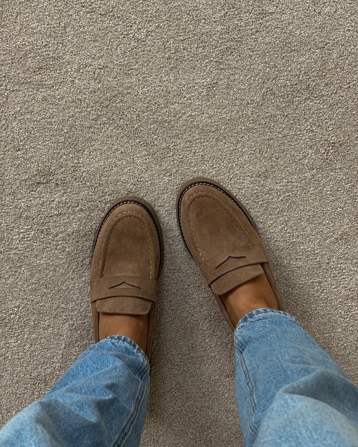 Tan suede loafers worn with light blue jeans on a beige carpet.