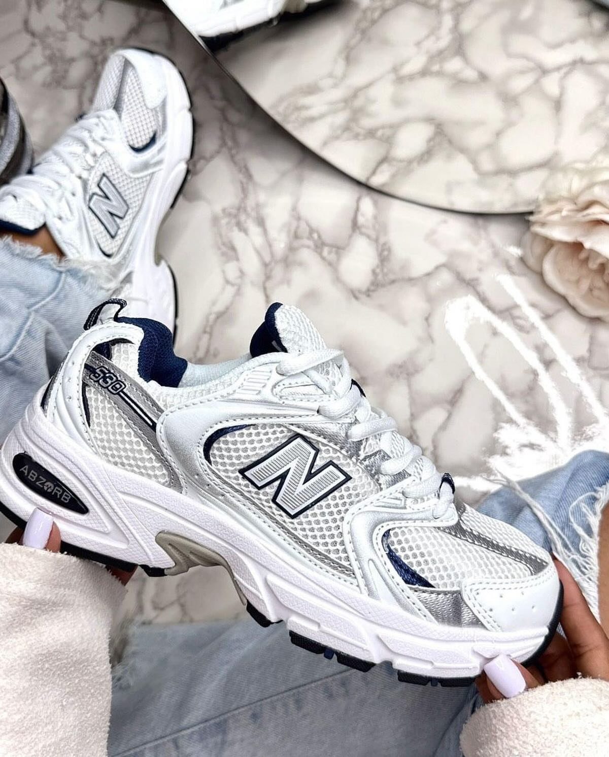 White New Balance sneakers with silver details held over light blue denim jeans.