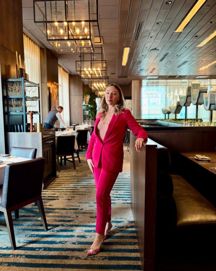 Woman in a bright pink pantsuit posing inside a modern upscale restaurant dining room