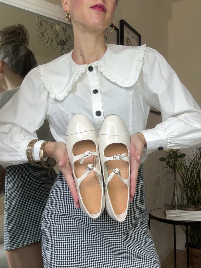 White patent leather Mary Jane flats held by a person in a white blouse.