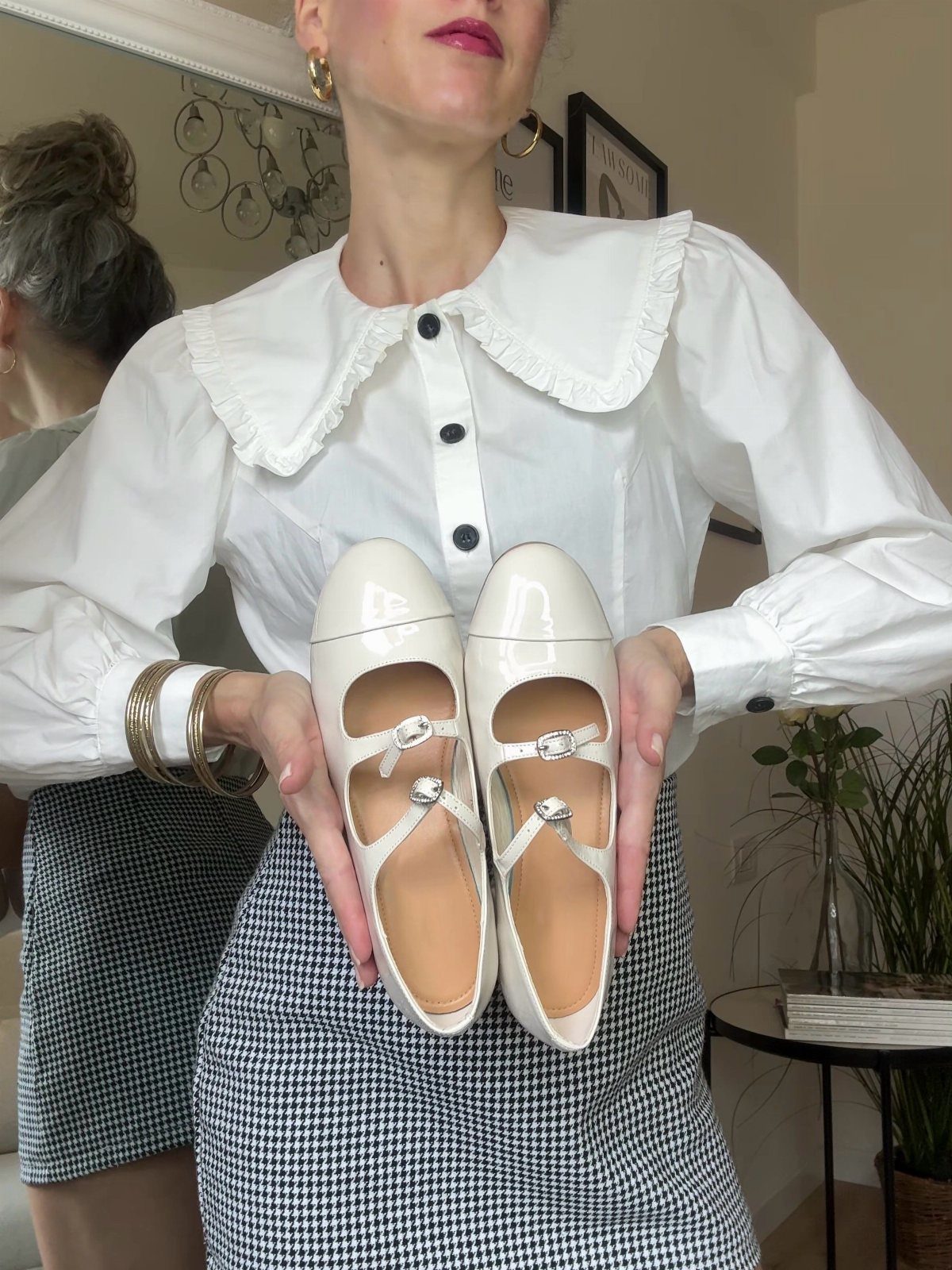 White patent leather Mary Jane flats held by a person in a white blouse.