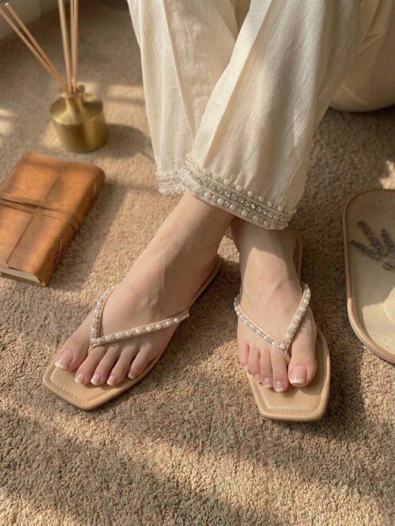 Beige square-toe flip flops with pearl-embellished straps worn with white pearl-trimmed trousers.
