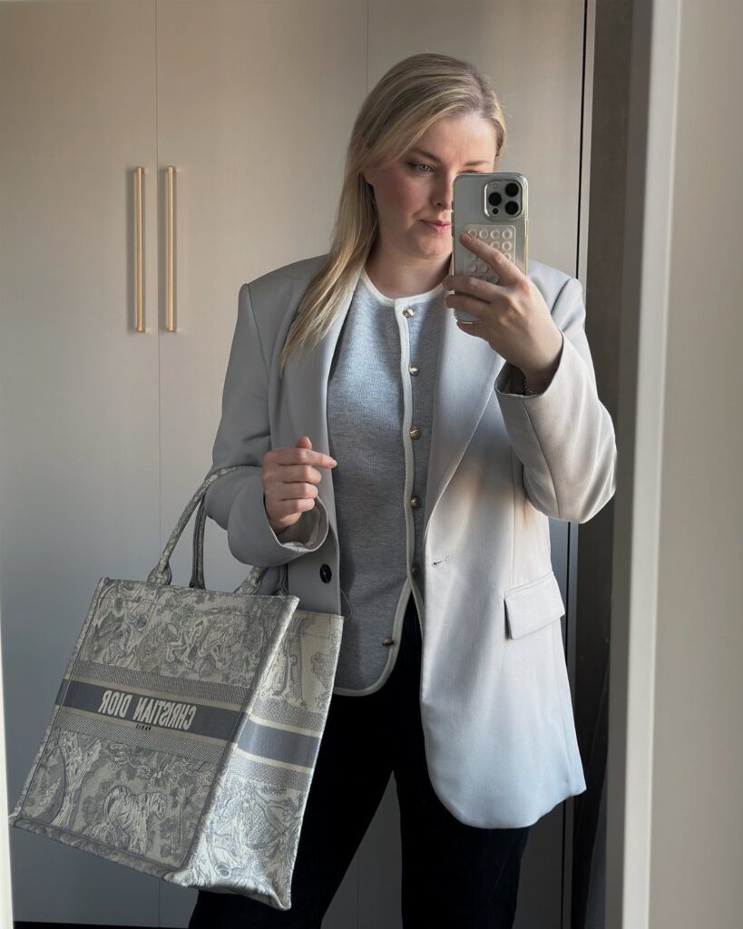 Woman in a grey blazer and black trousers holding a Dior tote bag.