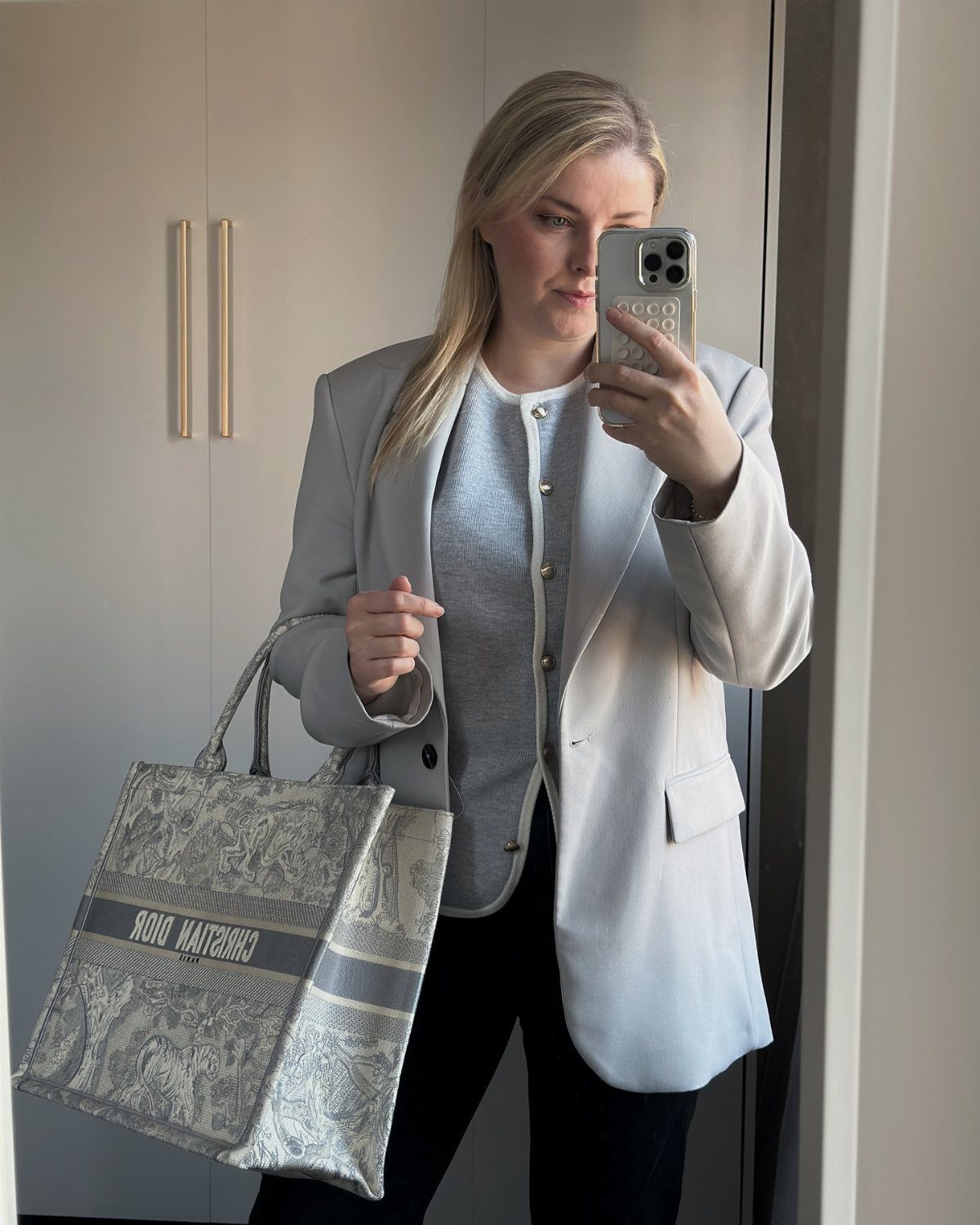 Woman in a grey blazer and black trousers holding a Dior tote bag.